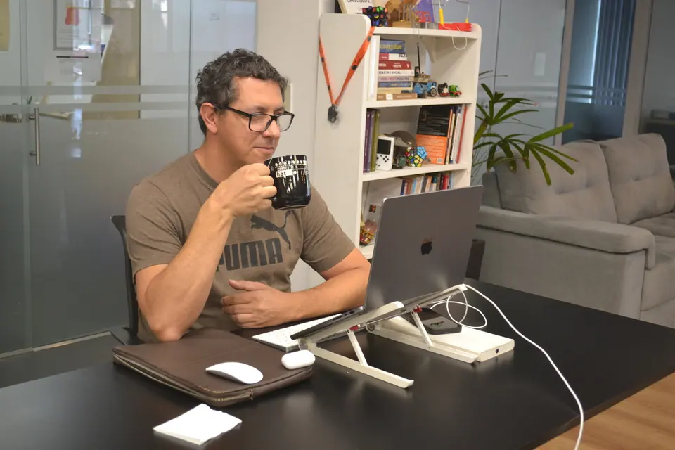 Persona tomando café frente a su notebook, con la biblioteca y los sillones detrás