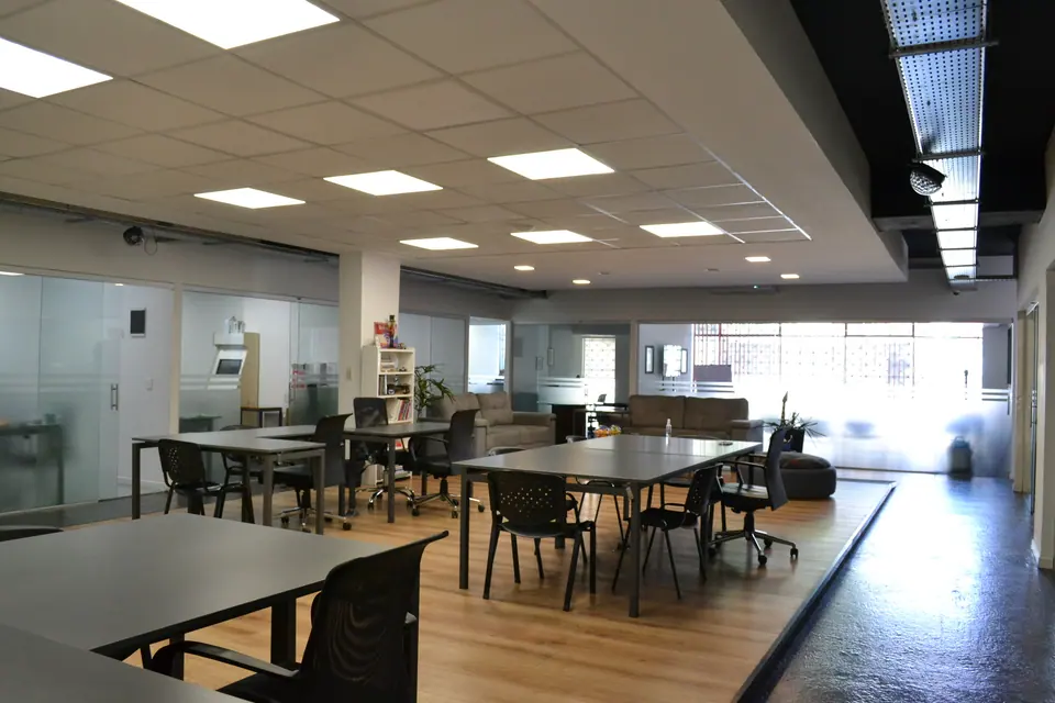 Vista amplia del espacio de cowork con mesas compartidas, piso de madera y oficinas vidriadas al fondo